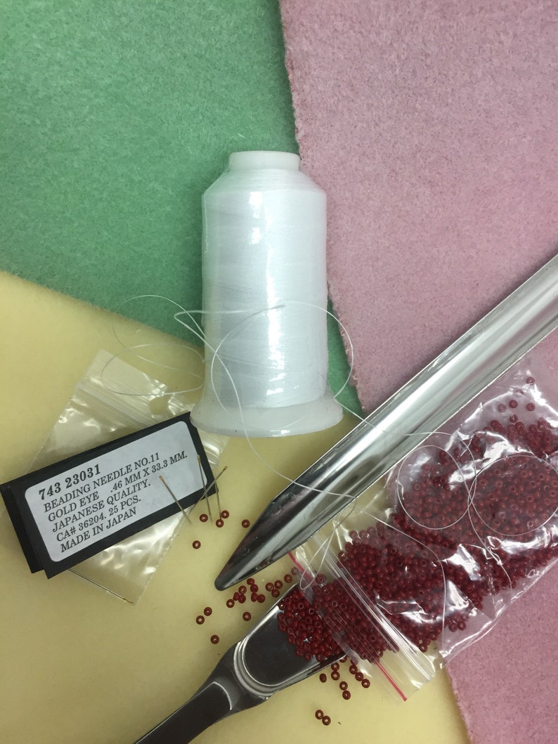 May include: A close-up of beading supplies including a spool of white thread, a metal beading tool, a small bag of red beads, and a package of beading needles labelled "743 23031 Beading Needle No. 11 Gold Eye 46 mm x 33.3 mm. Japanese Quality. CA# 36204. 25 Pcs. Made in Japan."