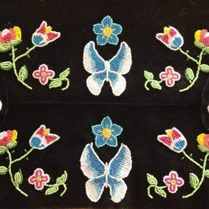 Könnte beinhalten: Schwarzer Stoff mit Perlenmotiven von Schmetterlingen und Blumen. Die Schmetterlinge haben weiße und blaue Flügel. Die Blumen sind rot, gelb und rosa. Die Perlenstiele und Blätter sind grün.