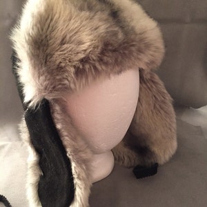 May include: A faux fur trapper hat with black suede ear flaps and a black chin strap. The hat is made of soft, fluffy faux fur and has a classic trapper hat style.