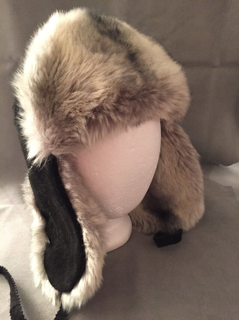May include: A faux fur trapper hat with black suede ear flaps and a black chin strap. The hat is made of soft, fluffy faux fur and has a classic trapper hat style.