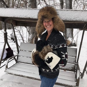 May include: A woman wearing a brown fur hat and a black and white patterned sweater is holding a pair of white mittens with a black bear silhouette on them. She is standing in a snowy backyard.