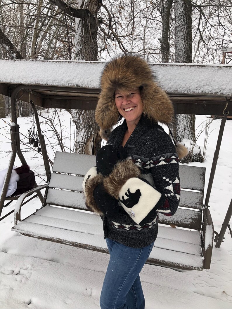 May include: A woman wearing a brown fur hat and a black and white patterned sweater is holding a pair of white mittens with a black bear silhouette on them. She is standing in a snowy backyard.