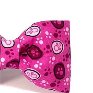 以下が含まれることがあります： 白と濃いピンクの足跡と足の形をした円の模様が入ったピンク色の蝶ネクタイ。 蝶ネクタイは鮮やかなピンク色で、ペット用である可能性があります。