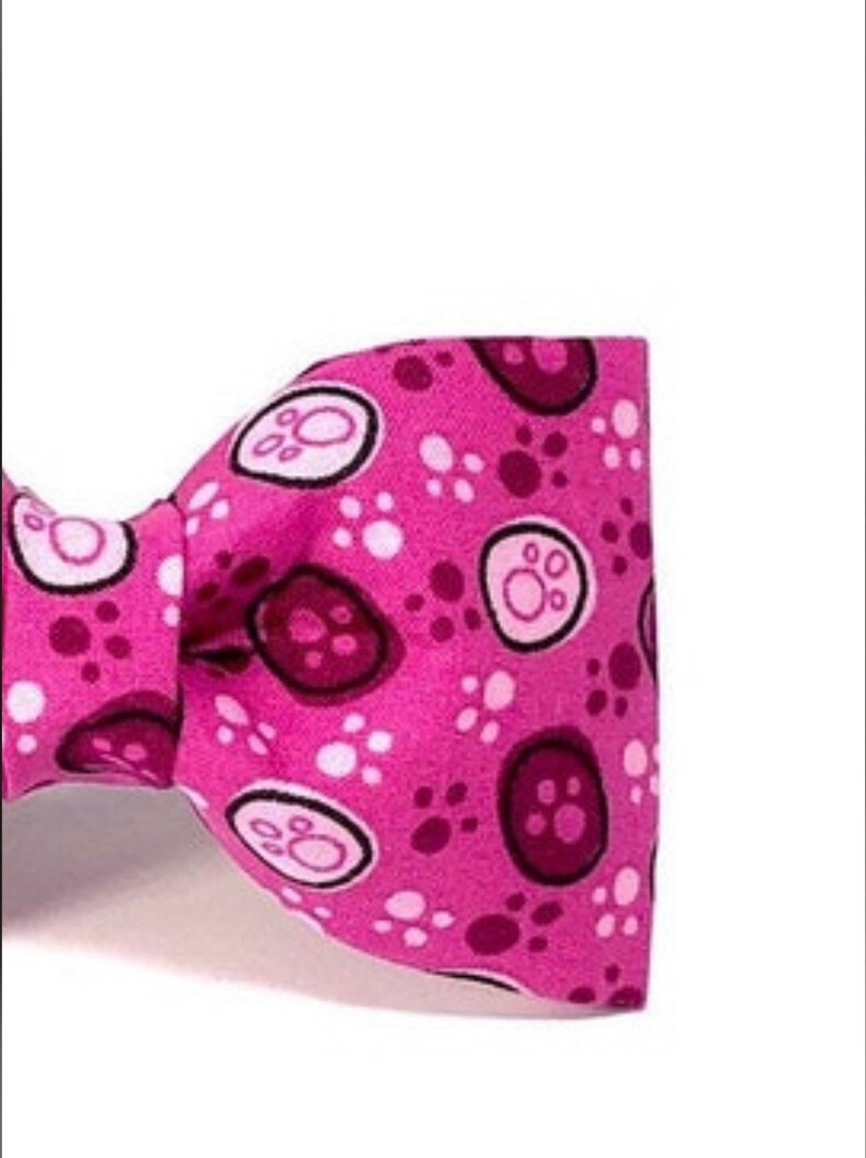 以下が含まれることがあります： 白と濃いピンクの足跡と足の形をした円の模様が入ったピンク色の蝶ネクタイ。 蝶ネクタイは鮮やかなピンク色で、ペット用である可能性があります。