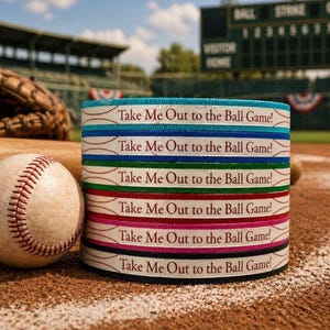 Op de afbeelding: Een honkbal en een stapel linten met de tekst "Take Me Out to the Ball Game!" staan op een honkbalveld. De linten zijn in verschillende kleuren, waaronder blauw, groen, rood en roze. Een honkbalhandschoen en een knuppel staan op de achtergrond.