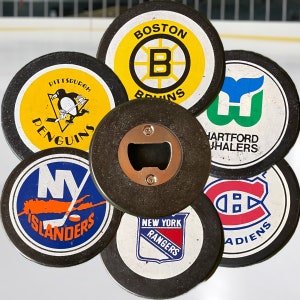 May include: A set of seven round, black and white hockey puck coasters with team logos. The logos include the Boston Bruins, Pittsburgh Penguins, New York Rangers, New York Islanders, Hartford Whalers, and Montreal Canadiens. The coasters are made of cork and have a bottle opener in the center.