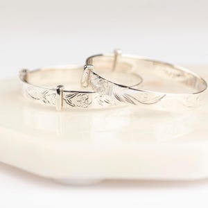 May include: Two silver-toned adjustable bangles featuring a delicate engraved floral pattern. Each bracelet has a smooth, polished finish and a sliding mechanism that allows for size adjustment, showcasing intricate botanical details along the band.