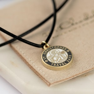 May include: A gold-colored pendant necklace with a black cord. The round pendant features a black border with the words "ST. CHRISTOPHER TRAVEL SAFE" and a white and gold image of St. Christopher. The necklace rests on a beige fabric.