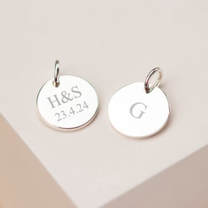 May include: Two silver round pendants with a loop at the top. One pendant is engraved with the initials "H&S" and the date "23.4.24". The other pendant has the letter "G" engraved on it.