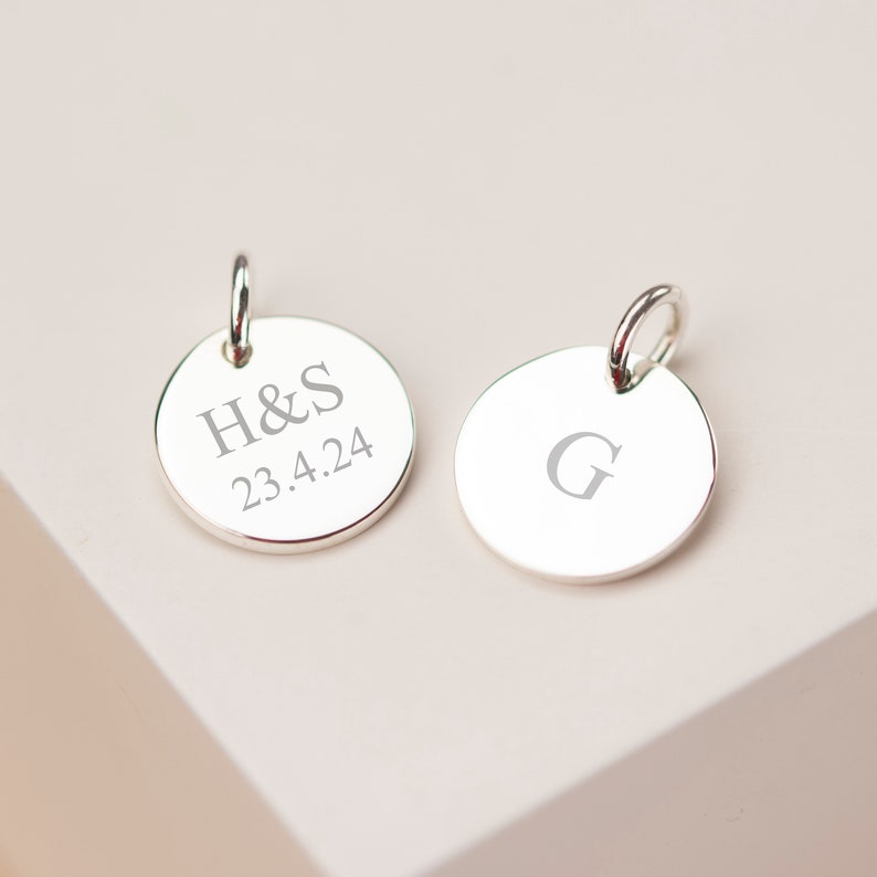 May include: Two silver round pendants with a loop at the top. One pendant is engraved with the initials "H&S" and the date "23.4.24". The other pendant has the letter "G" engraved on it.
