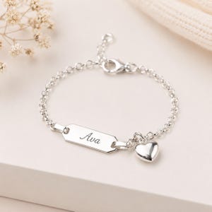 May include: A silver chain bracelet featuring a rectangular identification tag engraved with the name Ava in a cursive script font. The bracelet includes a small, smooth silver heart charm attached next to the tag and a lobster claw clasp.