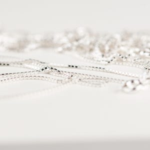 May include: Close-up of several silver box chain necklaces. The chains are scattered across a white surface, with a shallow depth of field. The links are square and reflective, catching the light. The chains are a simple, elegant jewelry item.