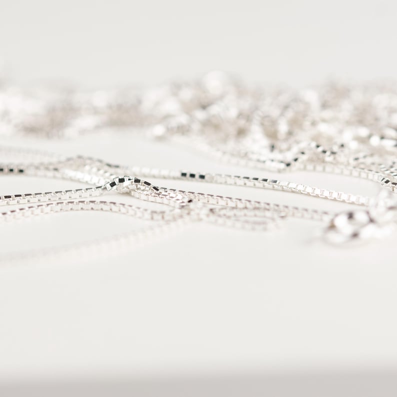 May include: Close-up of several silver box chain necklaces. The chains are scattered across a white surface, with a shallow depth of field. The links are square and reflective, catching the light. The chains are a simple, elegant jewelry item.