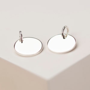 May include: Two silver-colored oval-shaped earrings with a polished finish. Each earring has a small loop at the top for attachment. The earrings are displayed on a white surface, showcasing their reflective quality.