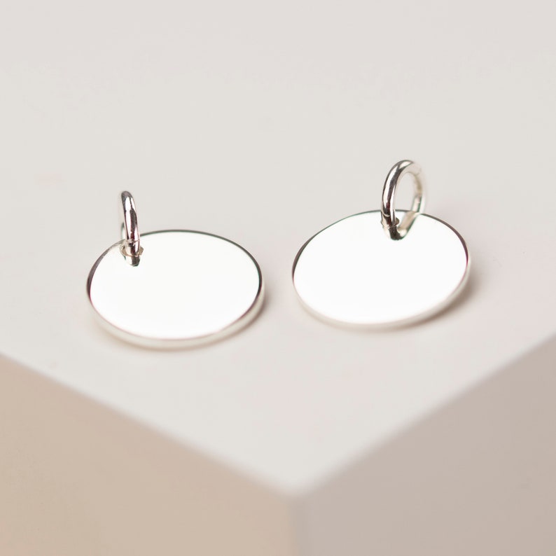 May include: Two silver-colored oval-shaped earrings with a polished finish. Each earring has a small loop at the top for attachment. The earrings are displayed on a white surface, showcasing their reflective quality.