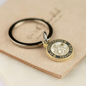 May include: A gold-colored keychain with a round pendant featuring the text "ST. CHRISTOPHER TRAVEL SAFE." The pendant has a black border and a white center with a detailed illustration. The keychain is on a beige surface.