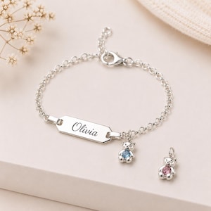 May include: A silver chain bracelet featuring a rectangular ID tag engraved with the name Olivia in a script font. The bracelet includes a small silver bear charm with a blue crystal and a separate silver bear charm with a pink crystal.