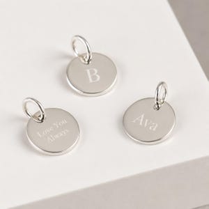 May include: Three circular silver-toned metal charms, each featuring a jump ring at the top. One charm is engraved with the letter B, another with the name Ava, and the third with the text Love You Always, all in a clean, sans-serif font style.