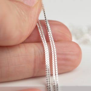 May include: Close-up of three delicate silver box chains. The chains are held by a hand, showcasing their fine, square links. The chains are displayed against a soft, white background, highlighting their reflective quality.