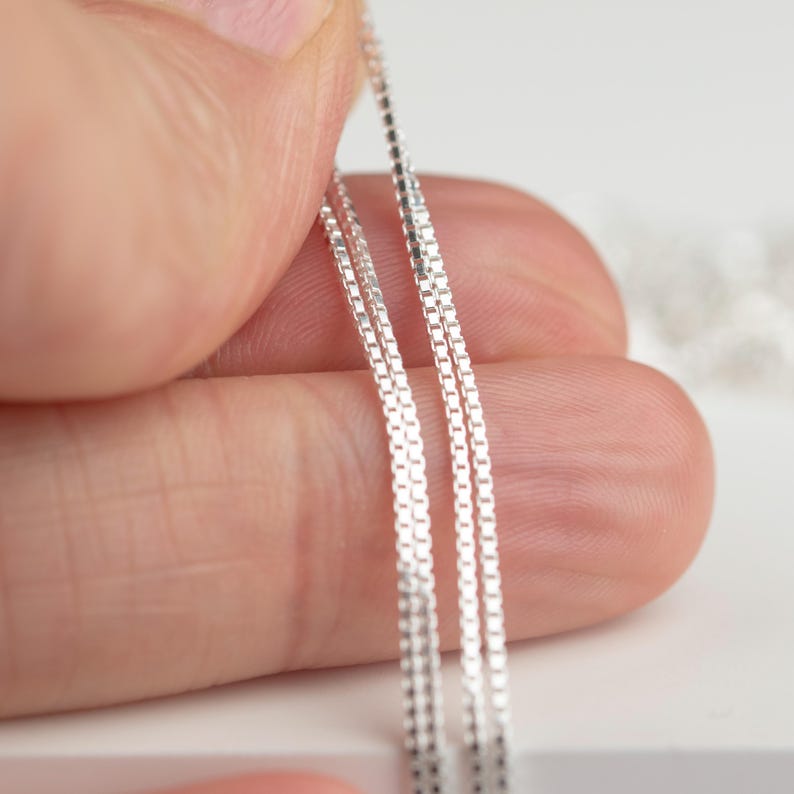 May include: Close-up of three delicate silver box chains. The chains are held by a hand, showcasing their fine, square links. The chains are displayed against a soft, white background, highlighting their reflective quality.