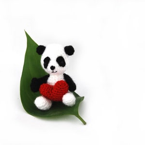 Puede incluir: Un pequeño panda de crochet sosteniendo un corazón rojo, sentado en una hoja verde.
