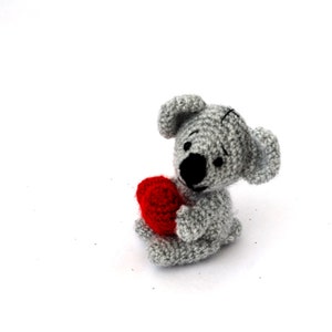 May include: A grey crocheted stuffed animal holding a red heart. The animal has a stitched-on nose and eyes.