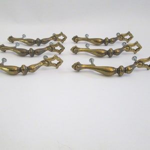 May include: Set of eight antique brass drawer pulls. Each pull features a decorative design with a central handle and ornate ends. The pulls are mounted with small screws, and are ideal for furniture restoration projects.
