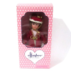 Puede incluir: Muñeca Effanbee vintage en una caja rosa con estampado de corazones. La muñeca viste un abrigo y sombrero rojos con ribetes blancos y detalles dorados. La cara de la muñeca es visible a través de la ventana de plástico transparente.
