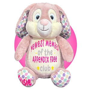 May include: A plush pink bunny with a cream-colored face and ears lined with colorful polka dots. The bunny has blue eyes and a pink nose. The text on the bunny reads "Newest Member of the Appendix Free Club" with a star and the name "Visie".