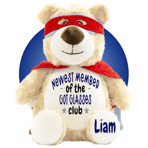 Può includere: Un orsacchiotto di peluche beige che indossa una maschera da supereroe rossa e un mantello rosso. Il petto dell'orso presenta il testo "NEWEST MEMBER of the GOT GLASSES club" in blu e nero, e il nome "Liam" è ricamato in blu sul piede destro.