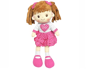 Könnte beinhalten: Eine Stoffpuppe mit braunen Wollhaaren, die zu Zöpfen mit rosa Schleifen gebunden sind. Die Puppe hat einen hellen Teint, rosige Wangen und trägt ein weißes Langarmshirt mit einem rosa Herz und dem Namen River sowie einen gepunkteten Rock und Schuhe.