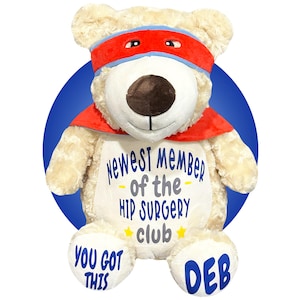 Puede incluir: Un oso de peluche crema con una máscara y capa de superhéroe rojas. El oso tiene el texto "Newest Member of the Hip Surgery Club" y "You Got This". El pie del oso tiene el nombre "Deb".