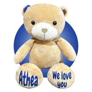 Könnte beinhalten: Ein kuscheliger, hellbrauner Teddybär mit einer blauen Schleife auf dem Kopf. Der Bär hat eine weiße Schnauze und eine herzförmige Nase. Auf den Füßen des Bären stehen die Worte "Athea" und "We love you" in Blau.