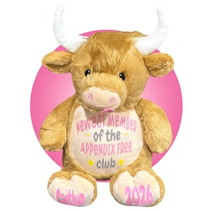 May include: A plush, light brown bull with white horns and a pink nose and tongue. The bull has the text "Newest Member of the Appendix Free Club" on its chest, with the name "Caitlyn" and the year "2026" on its feet.