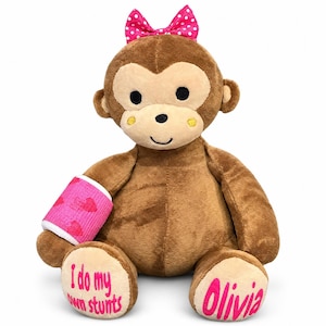 May include: A plush brown monkey toy featuring a pink polka dot bow on its head, a pink cast with red heart patterns on its right arm, and text on its feet reading I do my own stunts on the left and the name Olivia on the right in pink font.