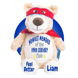 Puede incluir: Un oso de peluche con máscara y capa de superhéroe. El oso tiene bordado el texto "Newest Member of the Knee Surgery Club", "Feel Better" y "Liam". El oso es de color marrón claro con una cara blanca y un círculo azul detrás.