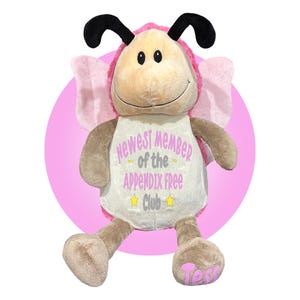 May include: A plush toy in the shape of a bee, with a tan body, pink wings, and black antennae. The toy has the text "Newest Member of the Appendix Free Club" on its chest. The name "Tess" is embroidered on one foot.