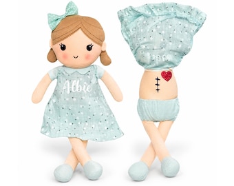 May include: A plush doll with brown yarn hair, a mint green bow, and a mint green floral dress featuring the name Albie. A second view shows the doll without the dress, revealing a red heart and a black stitched scar on the torso.