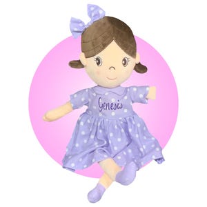 May include: A purple and white polka dot dress with a white collar and the name "Genesis" embroidered on the front. The doll has brown hair, a brown bow, and a light brown face.