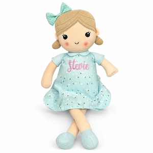 Puede incluir: Una muñeca de peluche con cabello de lana marrón claro en coletas y un lazo a juego. La muñeca viste un vestido azul claro con estampado floral blanco y el nombre Stevie bordado en rosa en el pecho, además de zapatos azul claro.