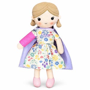 May include: A plush doll with light brown yarn hair, rosy cheeks, and a smiling face. The doll wears a cream-coloured dress with a colourful floral pattern, a purple cape, and pink shoes. A bright pink cast is visible on the doll's right arm.