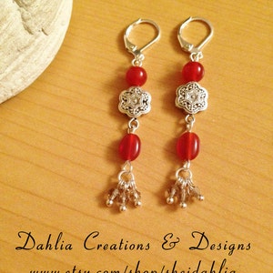 May include: A pair of silver earrings with red gemstone beads and a silver flower charm. The earrings have a dangling design with a cluster of small silver beads at the bottom.