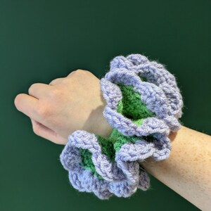 May include: Crocheted scrunchie in shades of lavender and green. The handmade hair accessory features a ruffled design, adding a touch of texture and color. This unique scrunchie is a stylish addition to any hair accessory collection.