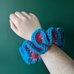 May include: A blue and pink crocheted hair scrunchie. The scrunchie is made with a thick yarn and has a ruffled texture.