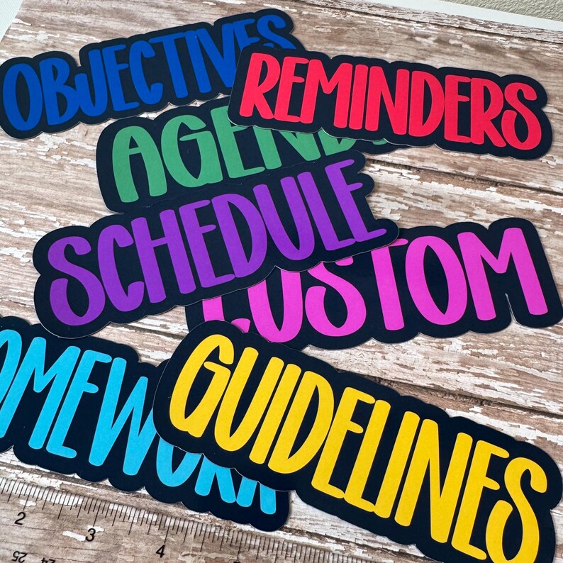 May include: A collection of colorful, die-cut labels with various words in bold, bubble letters. The labels include "OBJECTIVES," "REMINDERS," "AGENDA," "SCHEDULE," "CUSTOM," "HOMEWORK," and "GUIDELINES." Each label has a black border.