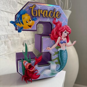 May include: A purple number 3 birthday decoration featuring Ariel, Flounder, and Sebastian from The Little Mermaid. The number has a mermaid scale pattern and the name "Gracie" in gold script.