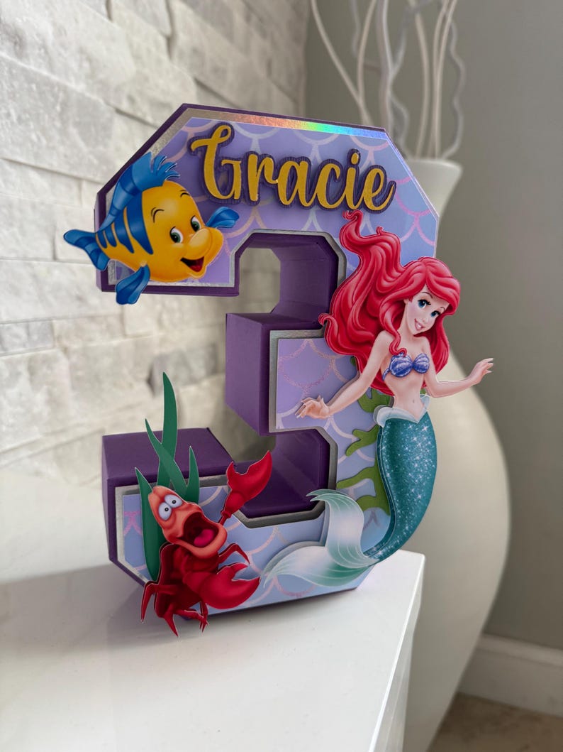 May include: A purple number 3 birthday decoration featuring Ariel, Flounder, and Sebastian from The Little Mermaid. The number has a mermaid scale pattern and the name "Gracie" in gold script.