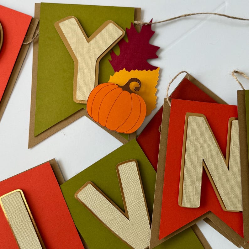 May include: A fall-themed banner with the word "Happy" spelled out in beige letters on green and red paper flags. Each letter is decorated with a gold border. The flags are strung together with twine. The "Y" flag has a small orange pumpkin and red and yellow leaves.