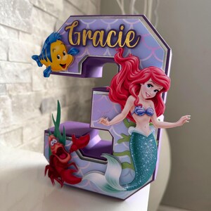 May include: A decorative number three featuring characters from The Little Mermaid. The number is purple and lavender with the name "Gracie" in gold script. Ariel, Flounder, and Sebastian are attached to the number.