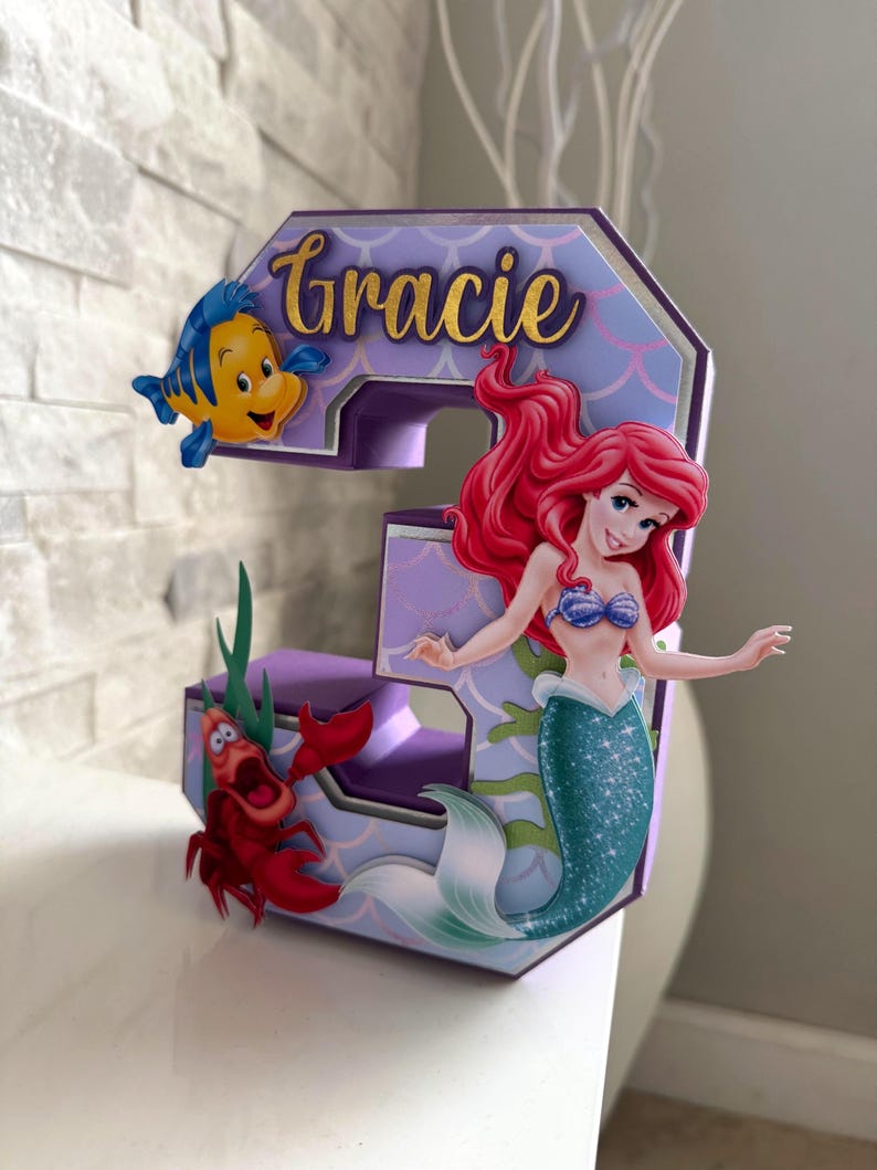 May include: A decorative number three featuring characters from The Little Mermaid. The number is purple and lavender with the name "Gracie" in gold script. Ariel, Flounder, and Sebastian are attached to the number.